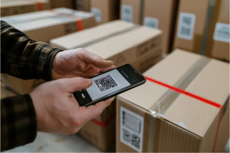 A person uses a smartphone to scan a QR code on a cardboard box in a warehouse, aiding in inventory management and asset tracking.