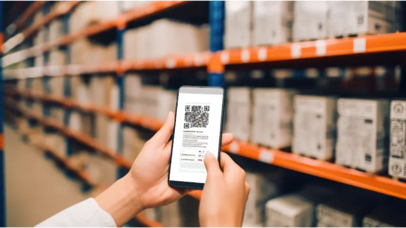 Person scanning a QR code on a smartphone in a warehouse with shelves of boxes in the background