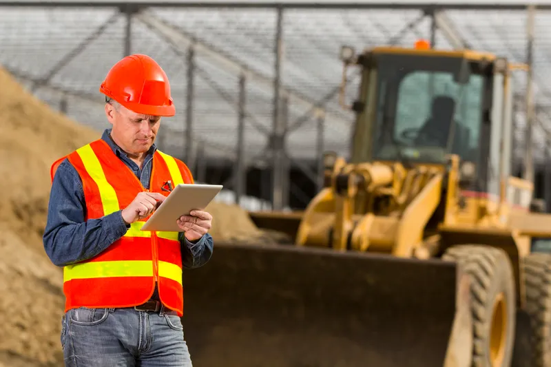 Construction worker in a red safety helmet and reflective vest using a tablet on a construction site, with a bulldozer in the background.