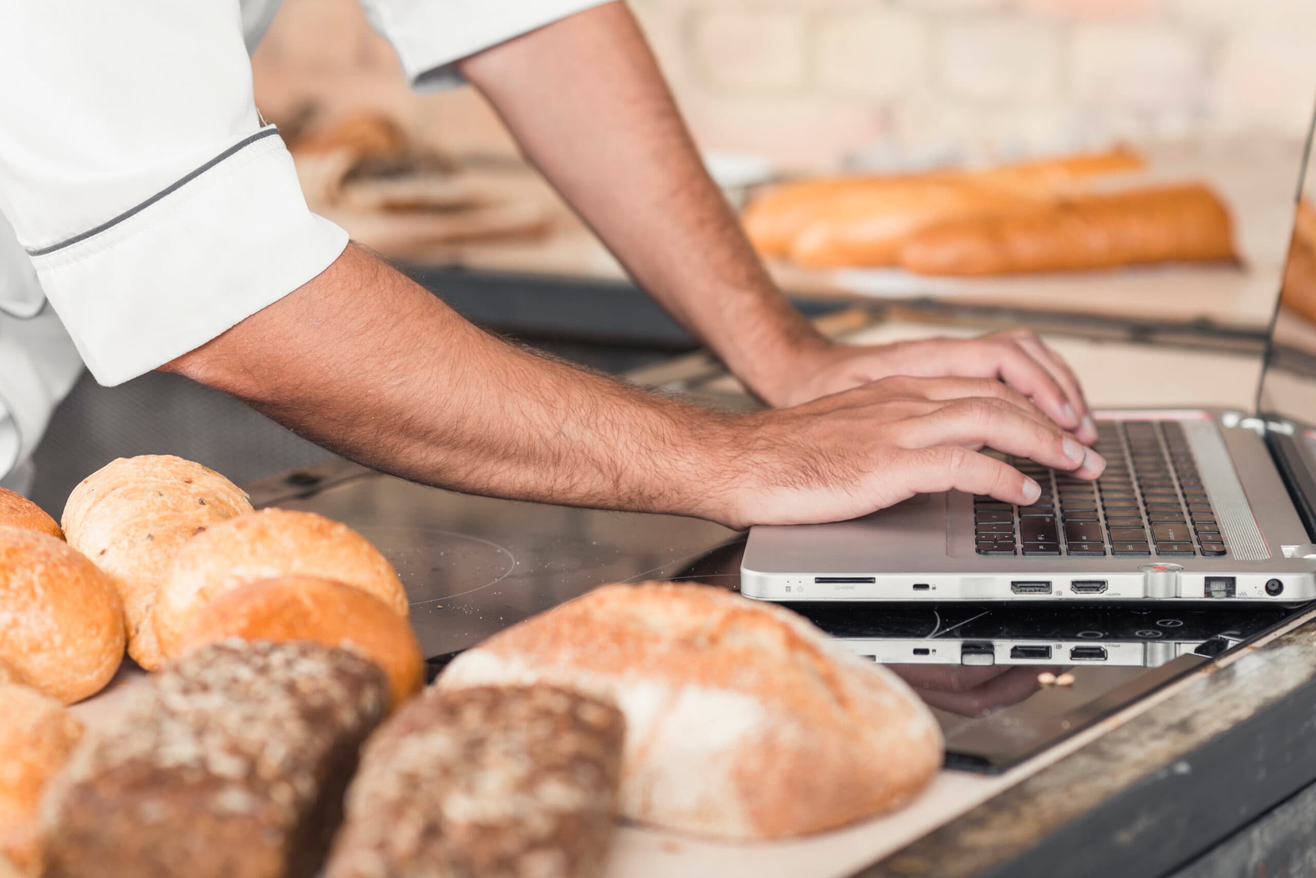 Close-up of a baker using a laptop on a kitchen worktop surrounded by freshly baked bread, highlighting the integration of technology in managing bakery operations.