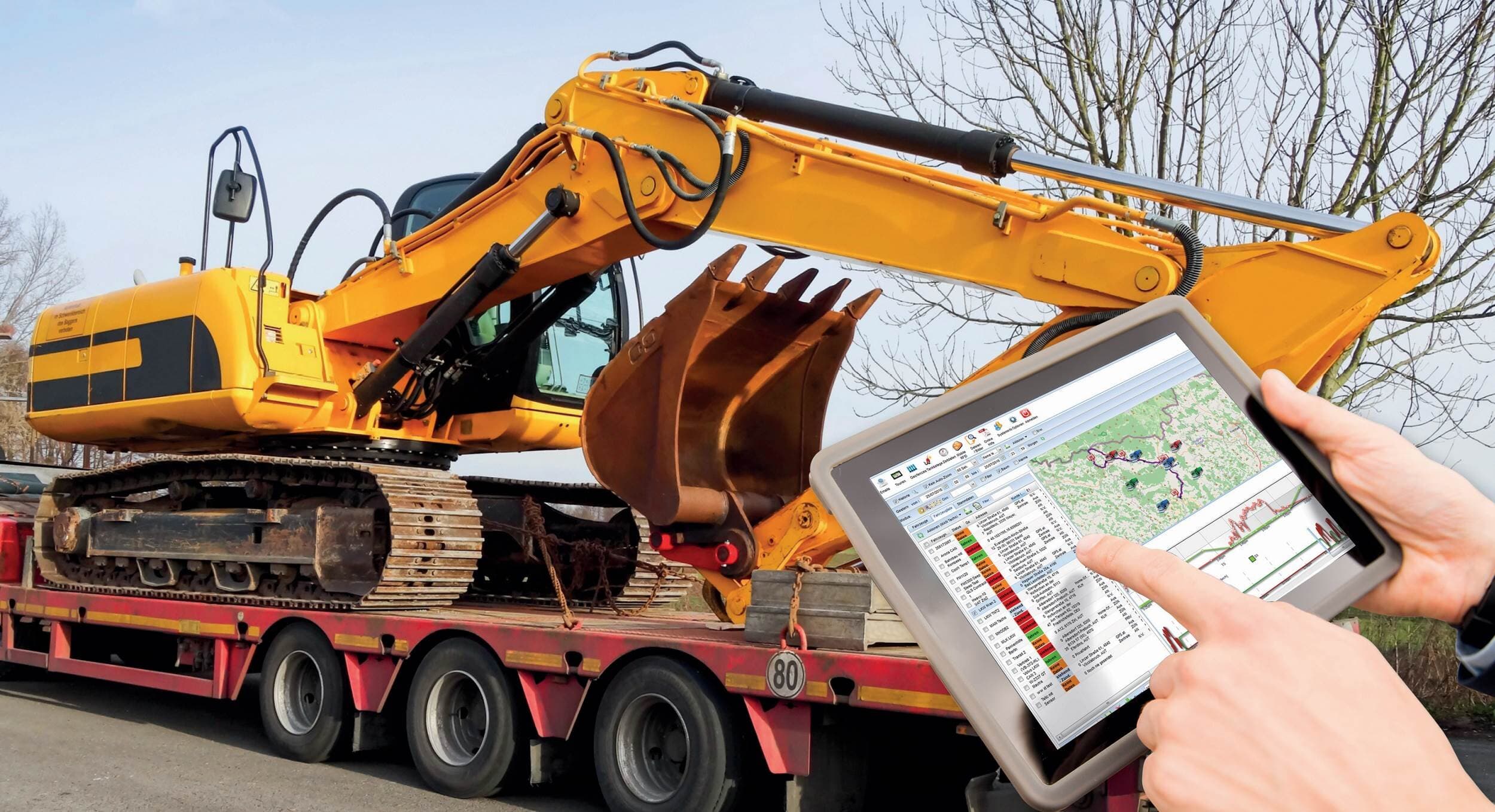Construction worker using a tablet to manage heavy machinery logistics, with a yellow excavator loaded on a flatbed trailer in the background.