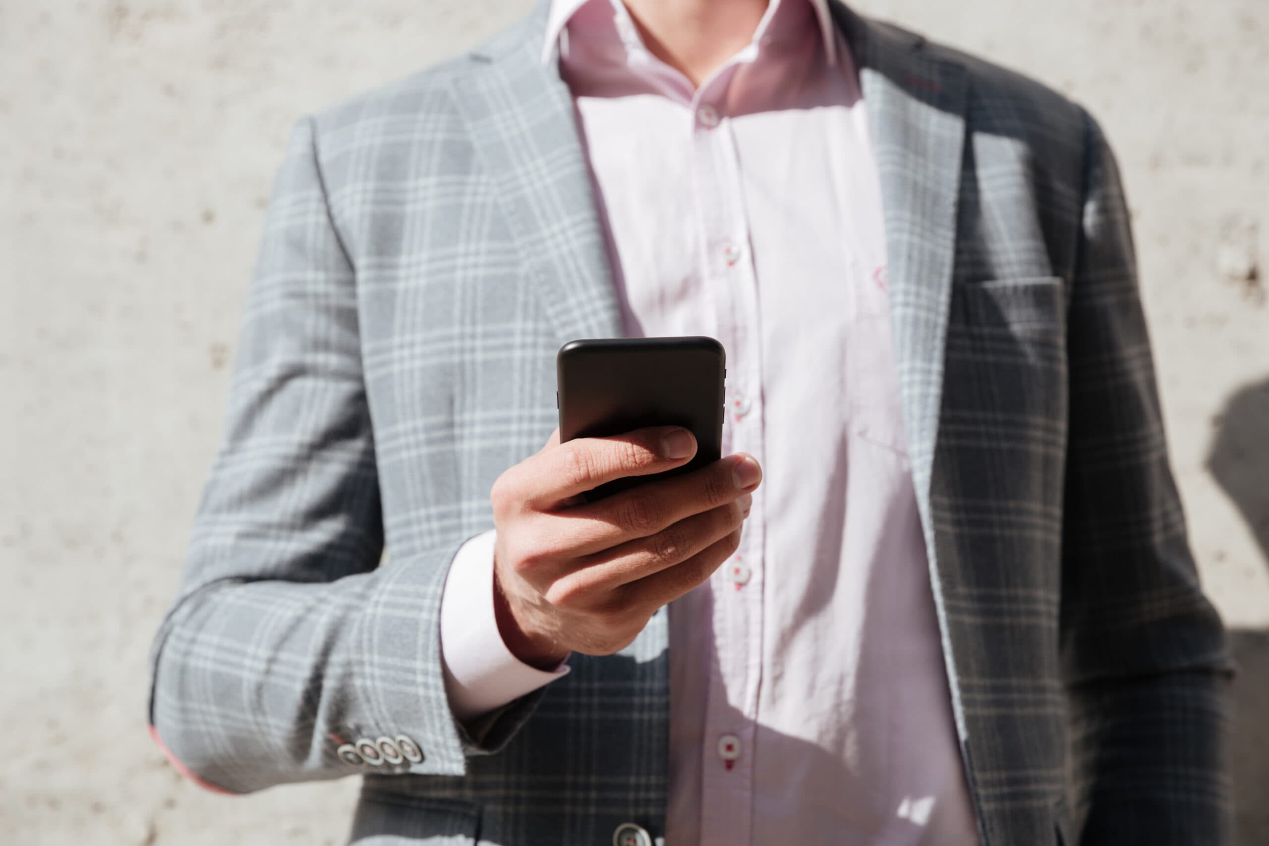 Businessman wearing a plaid suit jacket, holding a smartphone, with a neutral background in an outdoor setting.