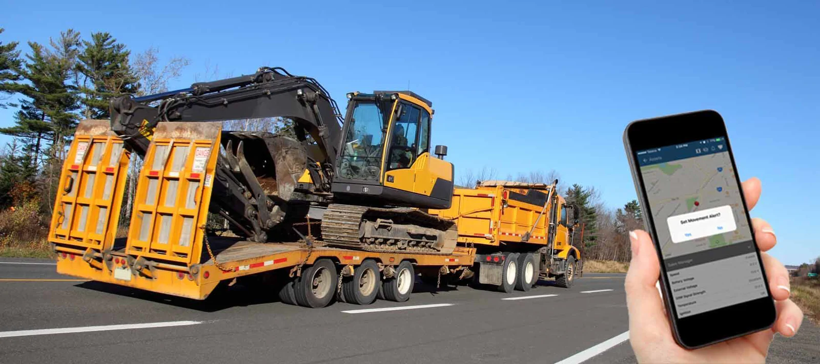 Heavy machinery being transported on a flatbed trailer, with a person using a smartphone to track and set movement alerts for the equipment.